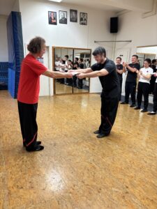 Sifu Bodo Seibold überreicht Lukas Fahsel mit gegenseitiger Verbeugung die Urkunde zum 2. Technikergrad im Wing Chun Kung Fu. Andere Schüler applaudieren im Hintergrund.