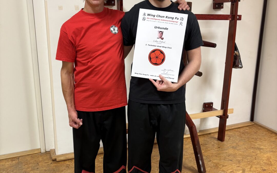 Lukas Fahsel steht neben Sifu Bodo Seibold und hält stolz seine Urkunde zum 2. Technikergrad im Wing Chun Kung Fu. Im Hintergrund steht eine traditionelle Wing-Chun-Holzpuppe.“