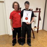 Lukas Fahsel steht neben Sifu Bodo Seibold und hält stolz seine Urkunde zum 2. Technikergrad im Wing Chun Kung Fu. Im Hintergrund steht eine traditionelle Wing-Chun-Holzpuppe.“